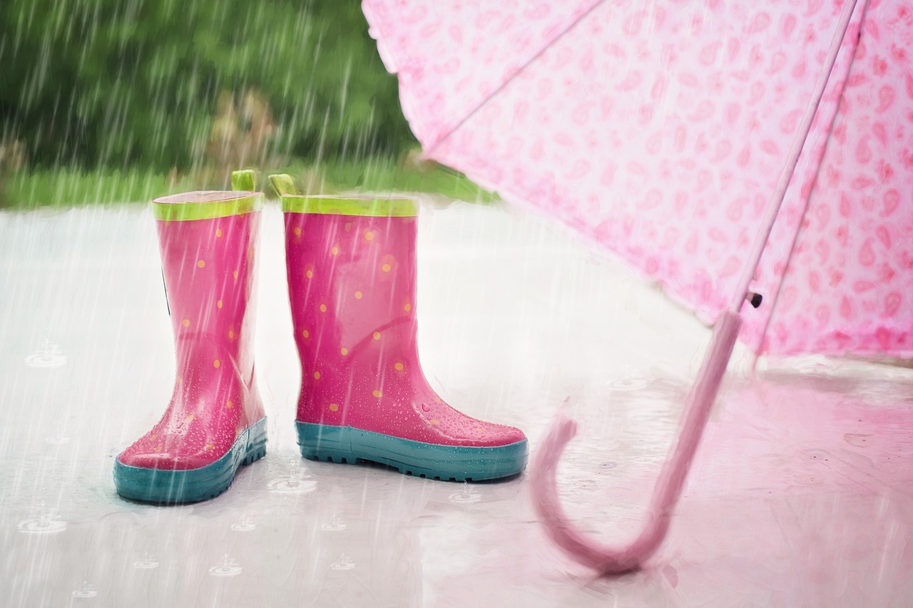 雨の中にピンクの長靴と傘が置いてある梅雨のイメージ